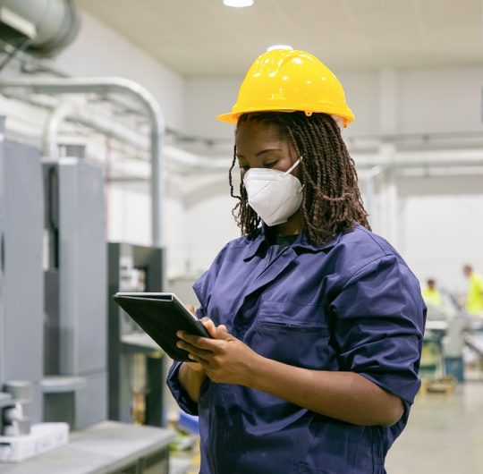 African American female factory worker in mask holding tablet. Focused content skilled woman working on plant and wearing protective helmet and uniform. Manufacture and digital technology concept